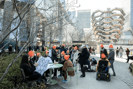 A group of about twenty people lounging in front of the Vessel, a building-sized, basket-like structure made of 154 interconnected stairways. The people are sitting, chatting, and reading. All wear neon orange Anti-Stairs Club Lounge beanies. 
