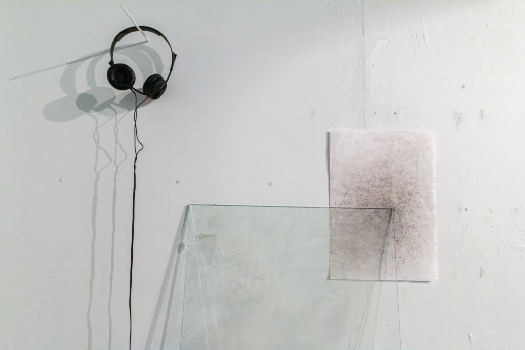 Black wired headphones hung on a white colored wall. A large panel of glass also leans against the white wall. Behind the upper right corner of the glass panel is a page covered with a variety of small, random marks in gray and reddish-brown.