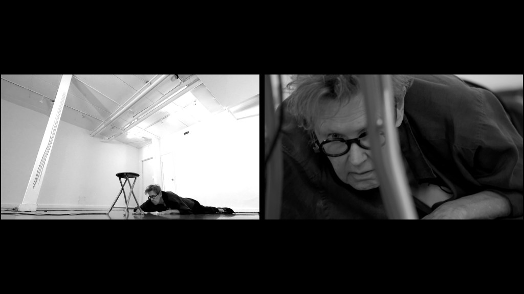 Two black and white images are side by side in a split screen.

In the image on the left, a woman lies on the floor looking up at a stool. There is a white column on the left with hand-drawn lines on it.

In the image on the right, a woman peers out over dark round glasses from under a stool. 
