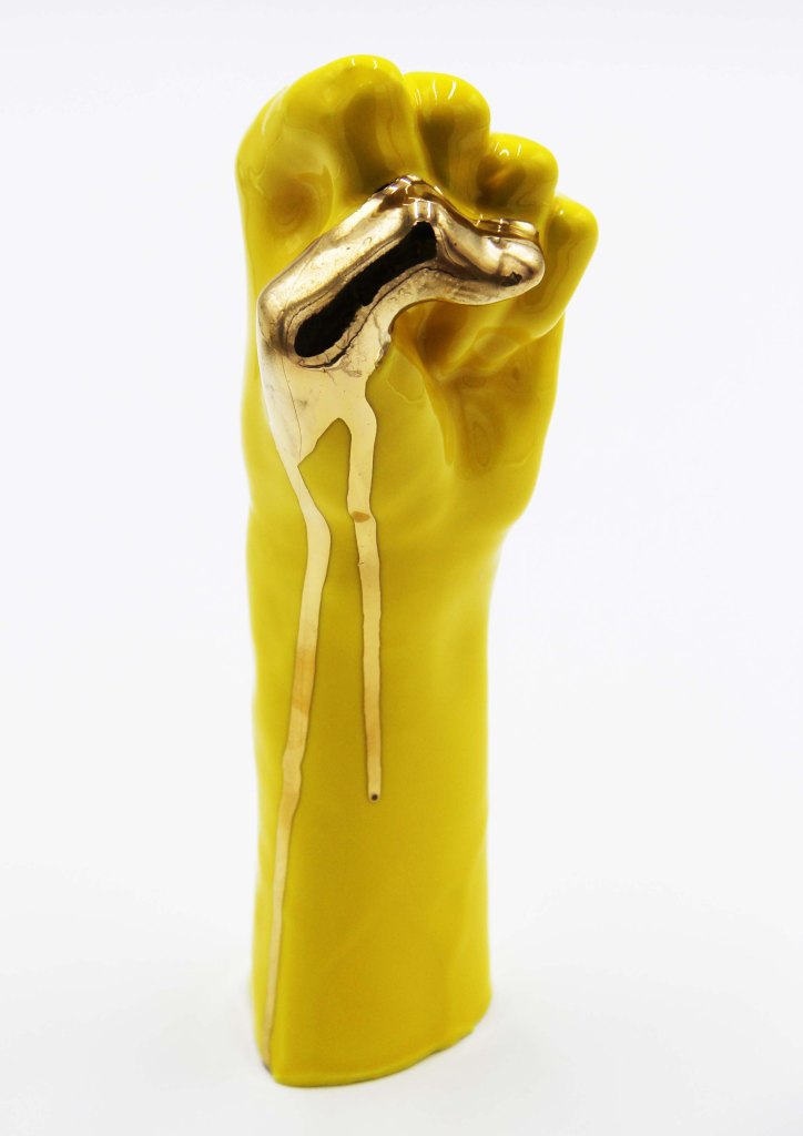 Yellow ceramic hand in an ASL S handshape. The thumb is painted gold with the paint dripping down the side of the wrist.