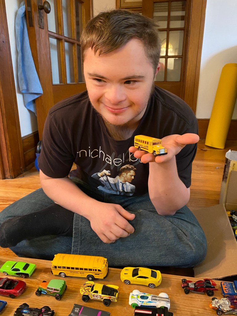 Logan holds up a yellow schoolbus toy while sitting cross legged at his table