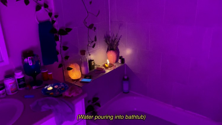 Film still. Purple light casts over the corner of a bathtub. Various medications, a salt lamp, a candle, and a smartphone with a glowing screen are along ledge and vanity behind the tub. Caption reads "Water pouring into bathtub".