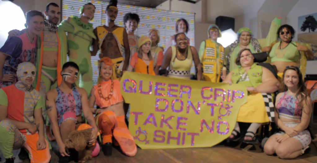 A group of people dressed in colorful clothing, holding a poster which says “Queer Crip Don’t Take No Shit”.