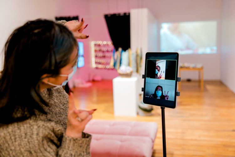 A person in an art gallery looks at an iPad mounted on a trolley. Two people in a video conference on the iPad gesture peace signs back. In the background a soft pink light and small white lights are through curtains to the left, a sculpture is in the centre, and a video is project on the wall on the right. 