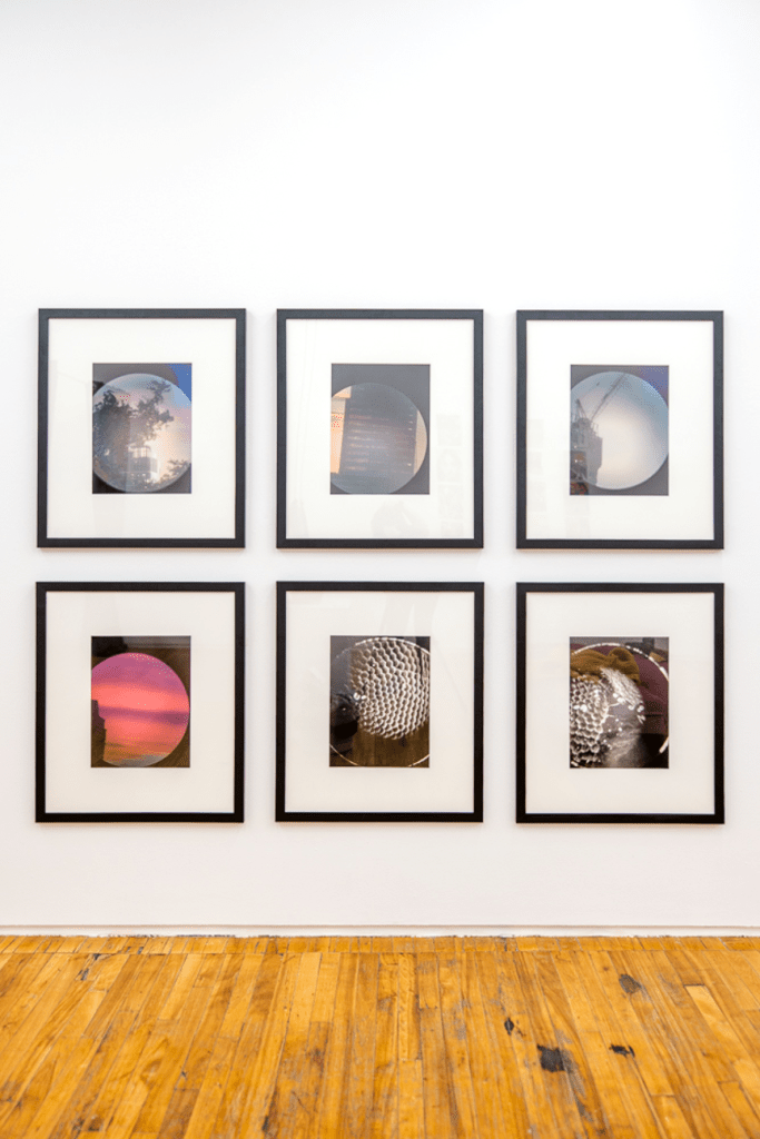6 framed photographs taken with a monocular lens are hung on a white wall. In the first row, the leftmost photograph is of a tree and a building in the background, taken on an iPhone 6 and shot through monocular. The adjacent photograph is an image of an apartment building taken on an iPhone 6, shot through monocular. The rightmost photograph is an image of a construction crane taken on an iPhone 6, shot through monocular. In the second row, the leftmost photograph is of a sunset sky with a building on the left taken on an iPhone 6, shot through monocular. The adjacent photograph depicts a rectangular image, a circle in the center. Blackness fills the space surrounding the rectangle’s perimeter. A blurry field emerges from the top center of the circle and fades to black. A bright white circle creates the circular plane. In the circle, an arrangement of smaller light gray circles glow in one big cluster. The cluster emerges from the blackness of the rest of the field. A white circle engulfs them with some room between it and the cluster. The right most photograph depicts a rectangular image, a circle in the center. Blackness fills the space surrounding the rectangle’s perimeter. A blurry field emerges from the bottom left side of the circle and fades to black. A bright white circle creates the circular plane. In the circle, an arrangement of smaller light gray circles glow in clusters. There is a stripey gray texture around them, almost like ridges.